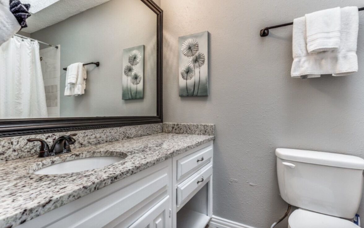 Un baño con lavamanos de encimera de granito, muebles de color blanco, inodoro blanco y dos obras de arte colgadas en la pared.