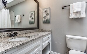 Un baño con lavamanos de encimera de granito, muebles de color blanco, inodoro blanco y dos obras de arte colgadas en la pared.