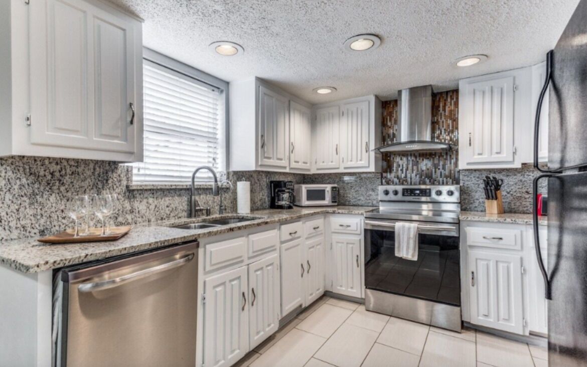 A modern kitchen with white cabinets, granite countertops, stainless steel appliances, and a window with blinds.