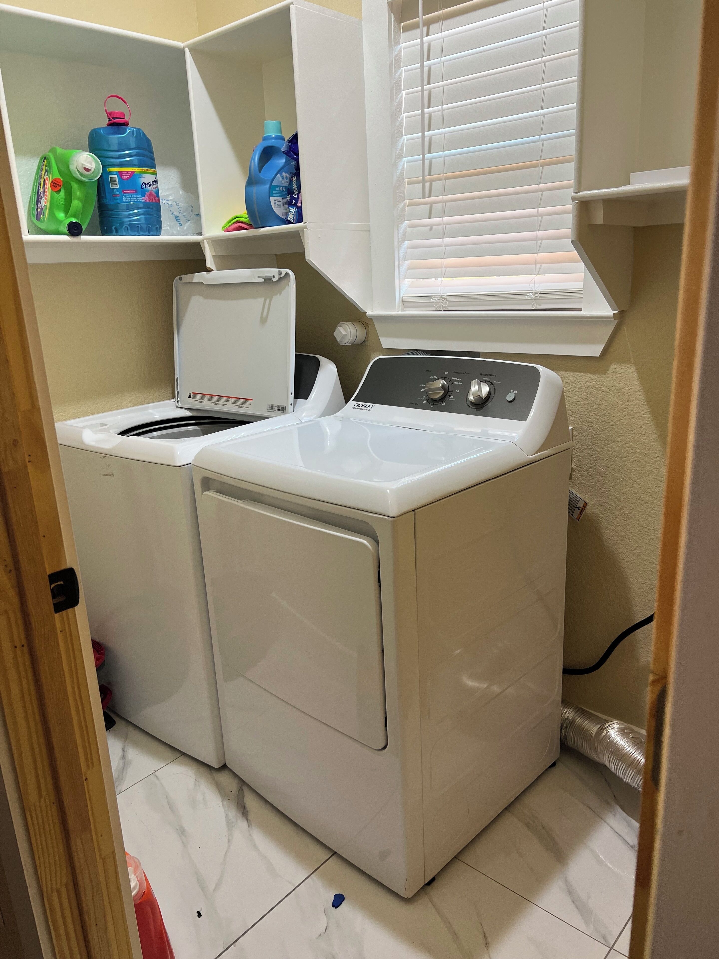 Área de lavanderia com máquina de lavar e secar de abertura frontal, prateleira com produtos de limpeza e janela com persianas.