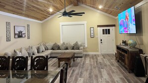 A living room with a wooden ceiling, a ceiling fan, a large TV, a sofa, a coffee table, and a dining table with chairs. - Home in Fort Worth (Fort Worth)