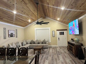 A living room with a wooden ceiling, a ceiling fan, a large TV, a sofa, a coffee table, and a dining table with chairs. - Home in Fort Worth (Fort Worth)
