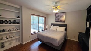 A bedroom with a bed, a ceiling fan, a window with blinds, a shelf with decorative items, and a painting on the wall. - Winding Hollow : Spacious 5-bedroom house in tranquil Arlington (Arlington)
