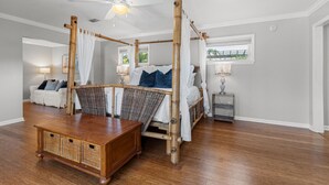 Ein Bett mit Baldachin, ein Holztisch mit Körben, ein Nachttisch mit Lampe und Blick ins Freie durch ein Fenster.