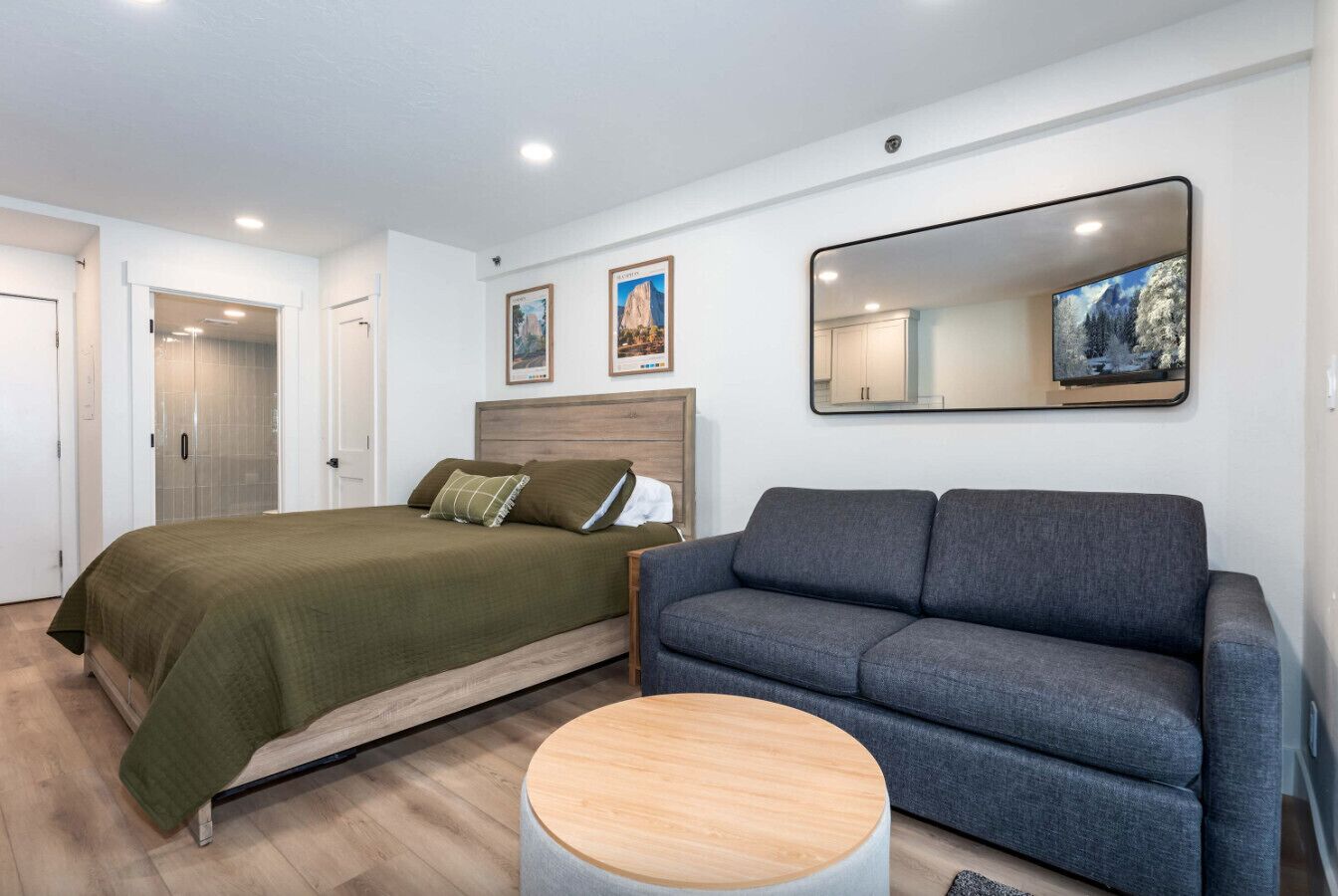 A modern bedroom with a bed, a grey sofa, a round wooden coffee table, and a large mirror on the wall.