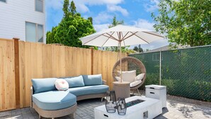 Eine Terrasse mit einer Eckcouch, einem runden Korbstuhl, einem weißen Couchtisch und einem großen Sonnenschirm.