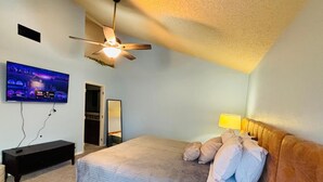 Een slaapkamer met een bed, een plafondventilator, een aan de muur bevestigde televisie, een bedlamp en een spiegel.