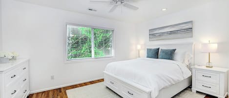 Une chambre à coucher avec un lit, des tables de chevet, un ventilateur de plafond, une fenêtre avec des stores et un tableau au mur.