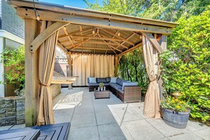 A covered outdoor seating area with a wooden pergola, cushioned seating, and a hanging light fixture.