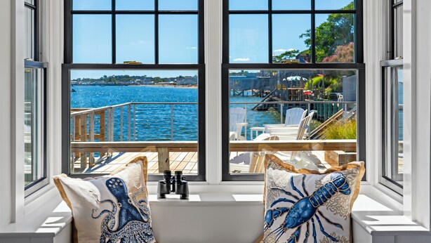 Asiento junto a la ventana con dos almohadas de pulpo, vista al agua y terraza con mobiliario exterior.