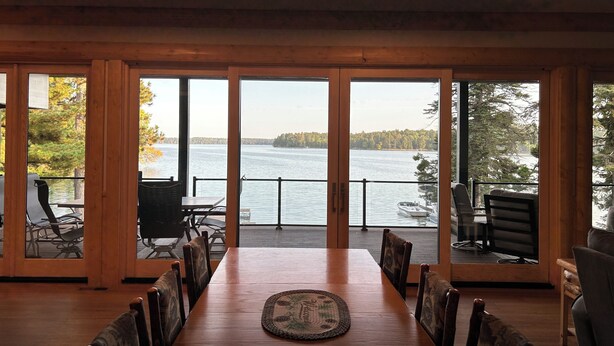 Un espace repas avec une grande table et des chaises en bois, de grandes fenêtres offrant une vue sur un lac et une forêt.