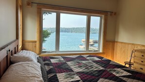 Une chambre avec une grande fenêtre donnant sur un lac, un lit recouvert d’une courtepointe et des lambris en bois.