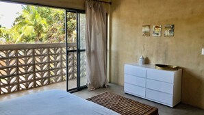 A bedroom with a bed, a white dresser, a small table with a decorative item, and a view of palm trees through a sliding glass door. -  Surrounded by beautiful views + 7 min. walk to the beach (Lo de Marcos)