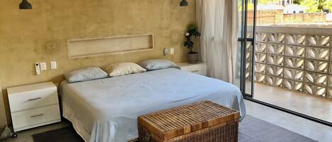 Un dormitorio con cama, mesitas de noche, un cofre de mimbre y vistas al verdor exterior.