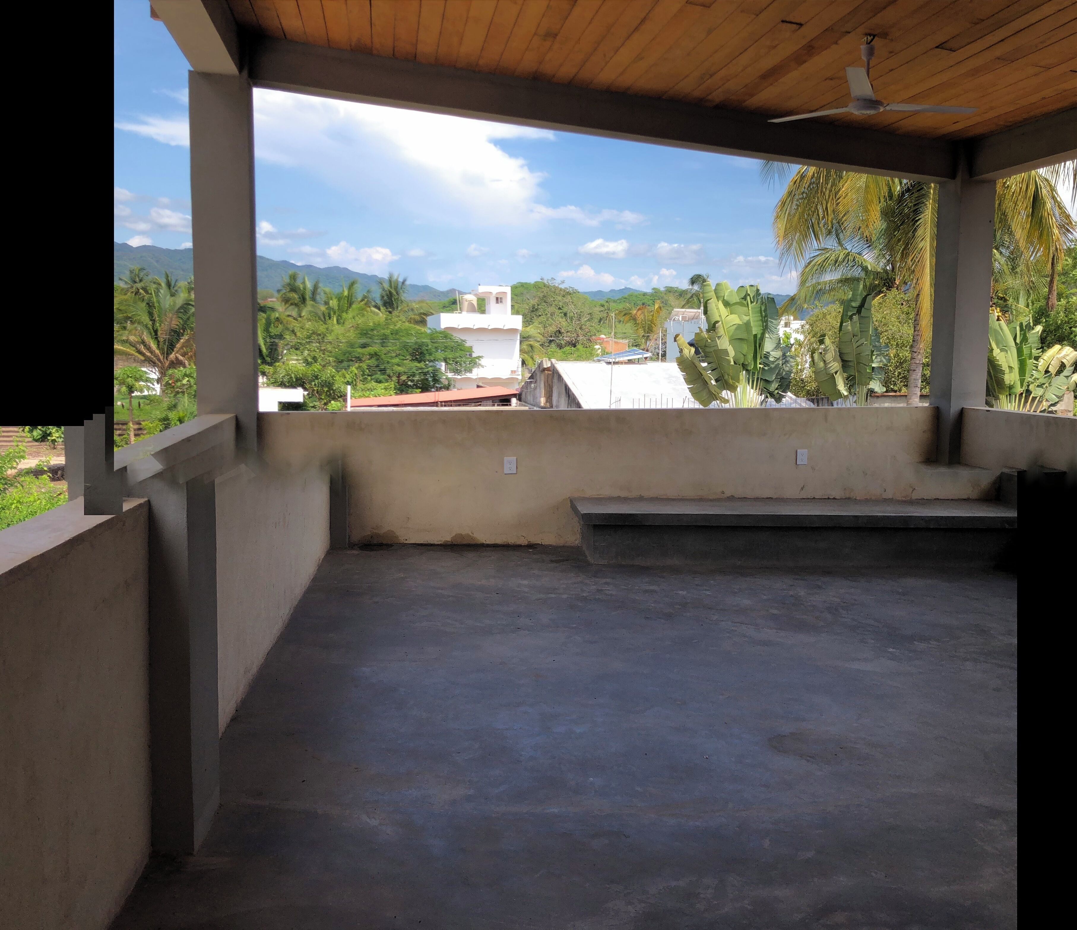 An empty covered outdoor area with concrete flooring, a ceiling fan, and a view of surrounding buildings and greenery.