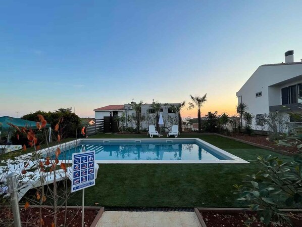 Une piscine avec deux chaises longues, un panneau indiquant les règles de la piscine, et une maison moderne en arrière-plan.