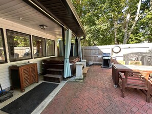 A covered patio with a brick floor, a wooden table and chairs, a grill, and a covered seating area.