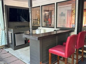 An outdoor kitchen area with a built-in grill, a refrigerator, and a bar counter with two red bar stools.