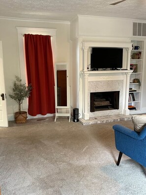 Een woonkamer met een open haard, een televisie, een blauwe fauteuil en een rood gordijn.