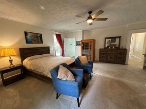 A bedroom with a bed, two armchairs, a dresser, a ceiling fan, and a painting on the wall.