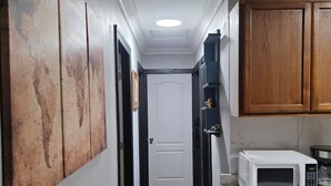A narrow hallway with wooden flooring, a white door at the end, and a microwave on a counter to the right.
