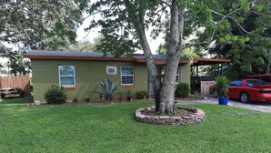 A house with a well-maintained lawn, a tree in the center, and a red car parked in the driveway.