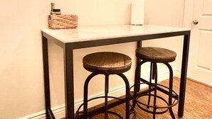 Ein kleiner Essbereich mit einem weißen Tisch und zwei Hocker, einer Wanddekoration mit Stadtansicht, einer Kerze und einem Korbwäscher.