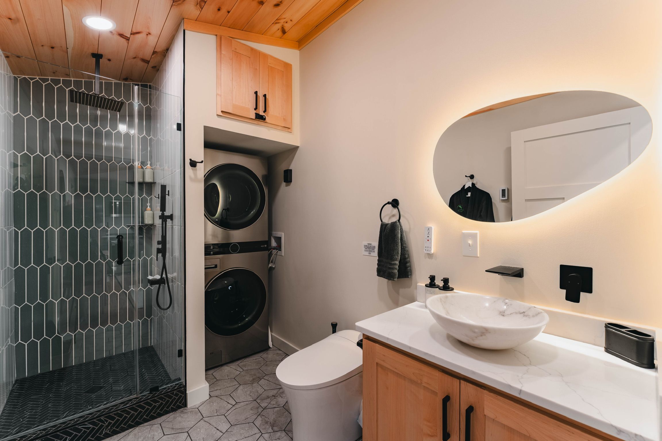 A modern bathroom with a glass shower enclosure, a built-in washing machine, a large oval mirror, and a white sink.