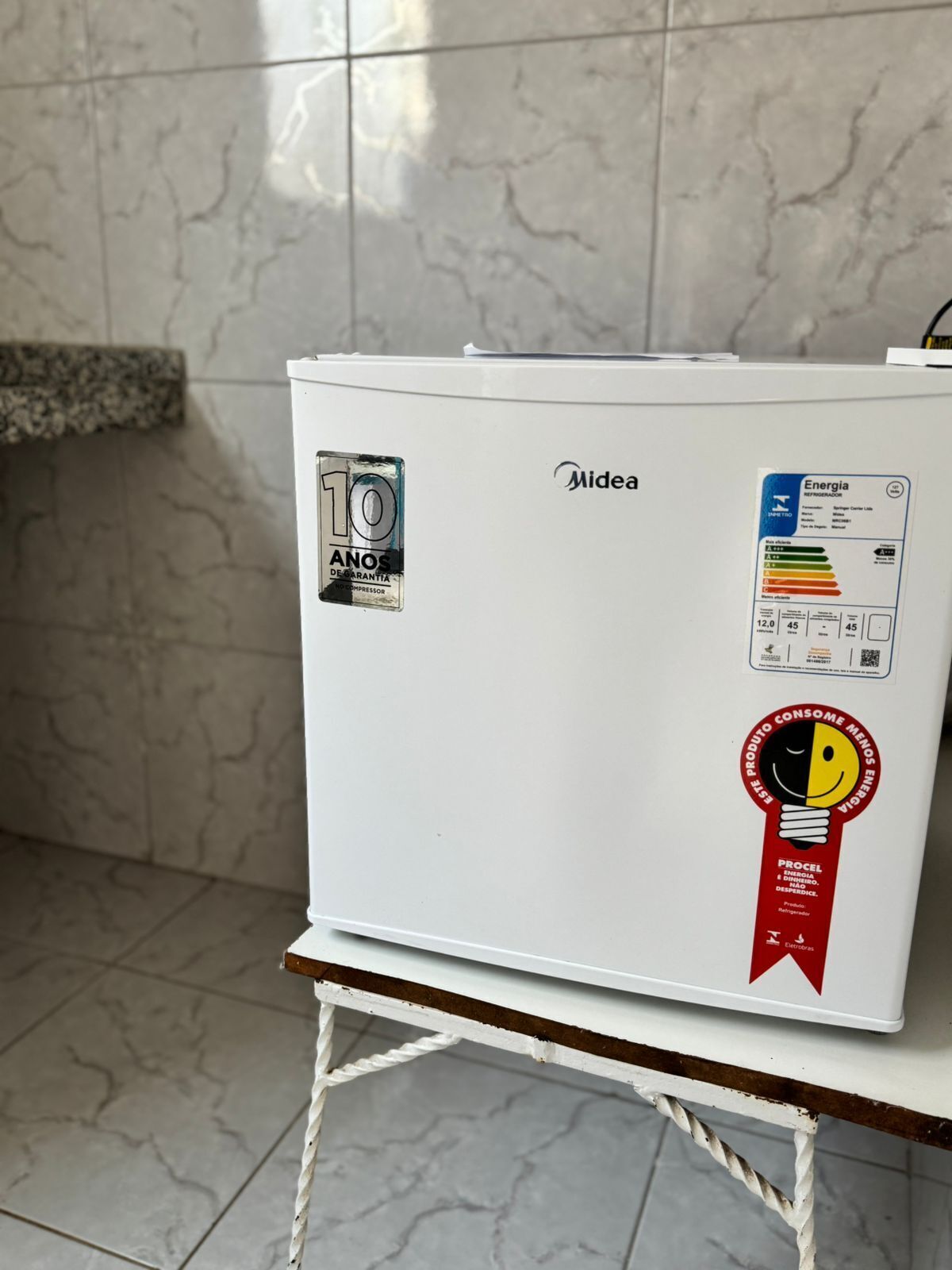 A white refrigerator with a 10-year warranty sticker, a sticker indicating it's energy efficient, and a red sticker with a light bulb and the text 'PROCEL'.