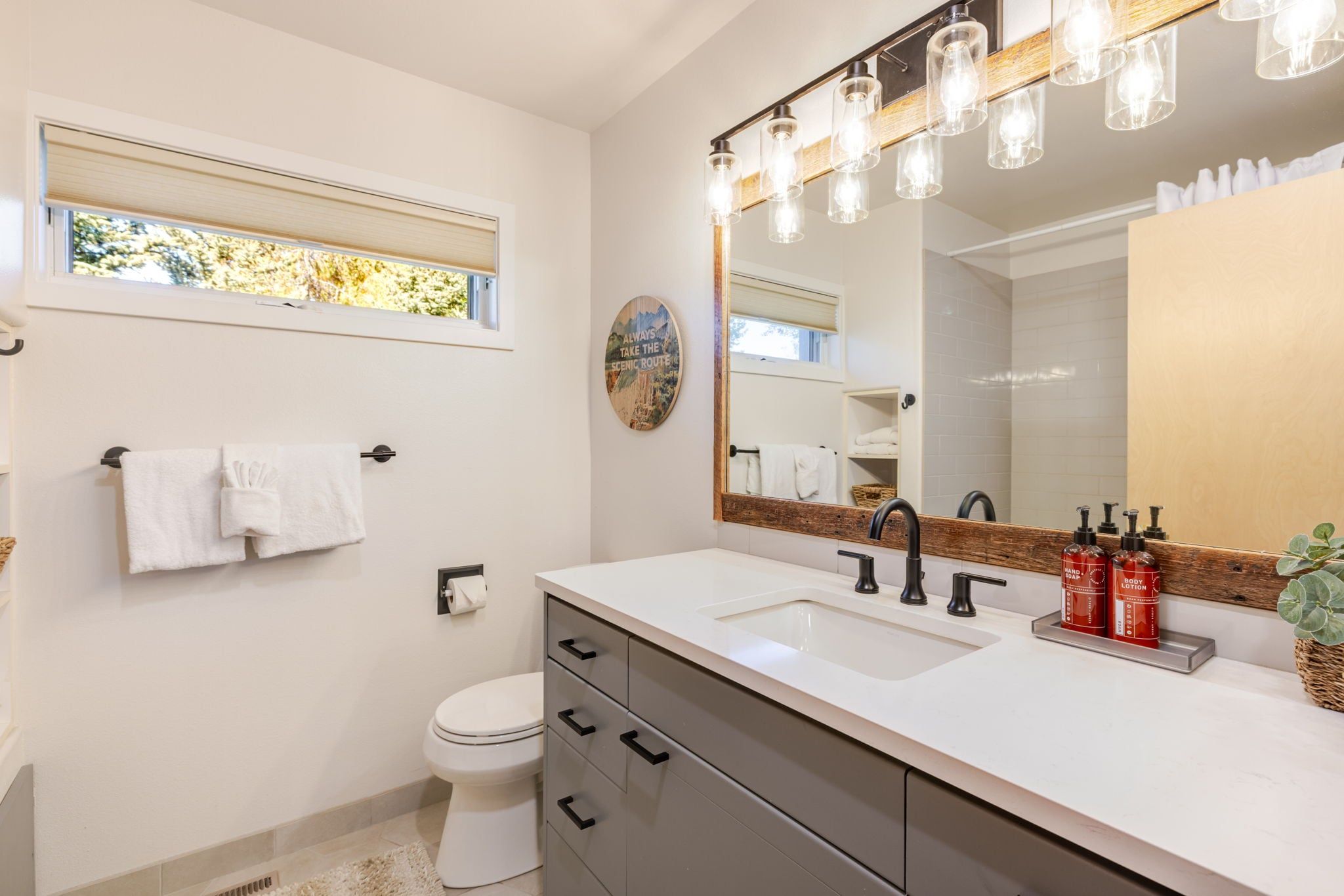 A modern bathroom with a large mirror, a window, a toilet, and a vanity with a sink and storage drawers.