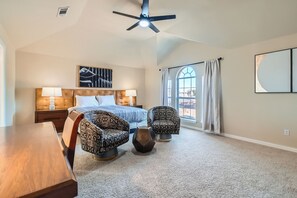 Quarto com cama, duas poltronas estampadas, mesa de madeira, ventilador de teto e uma janela grande com cortinas.