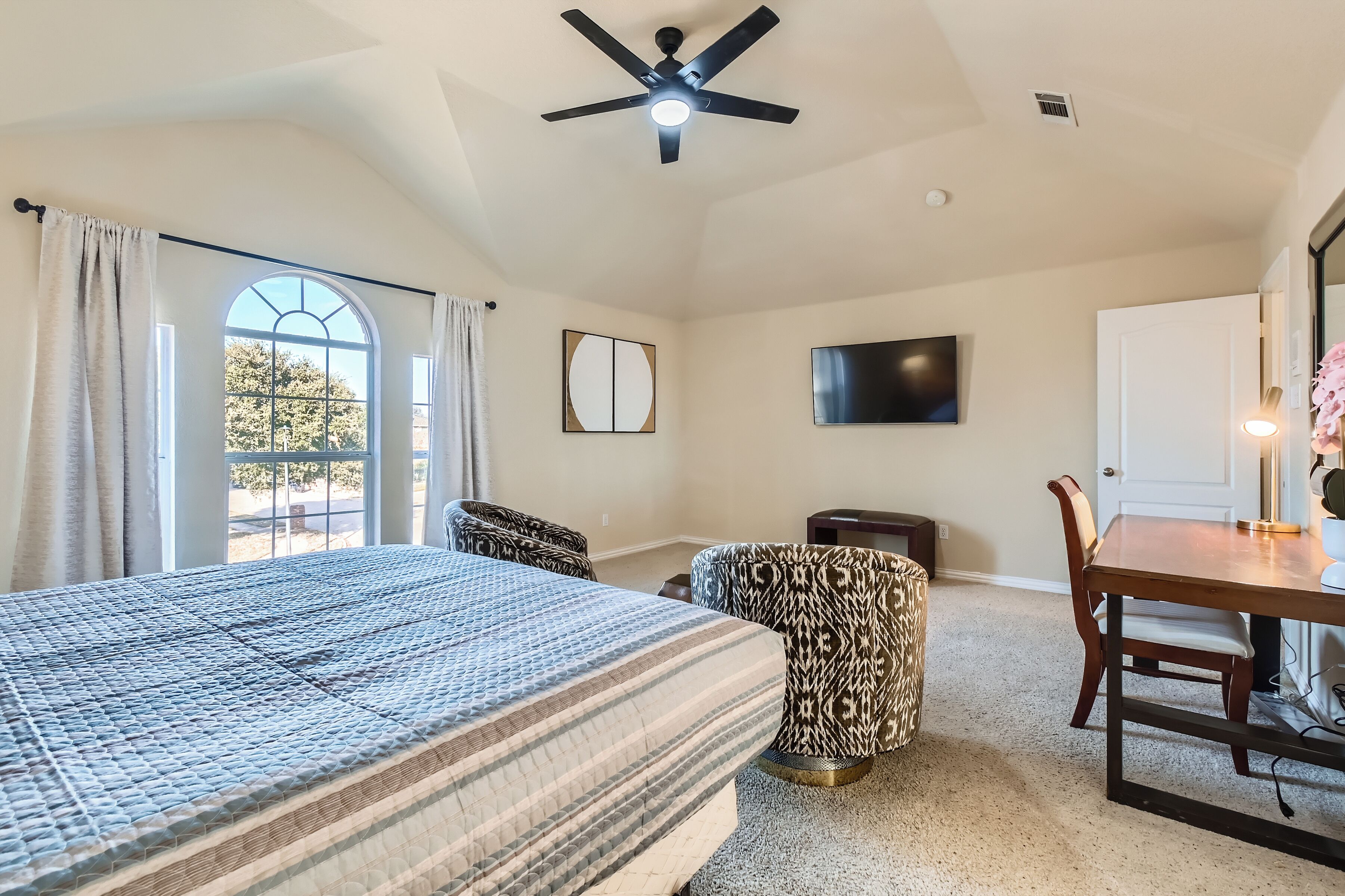 A bedroom with a bed, a desk, a chair, a television, and a window with curtains.