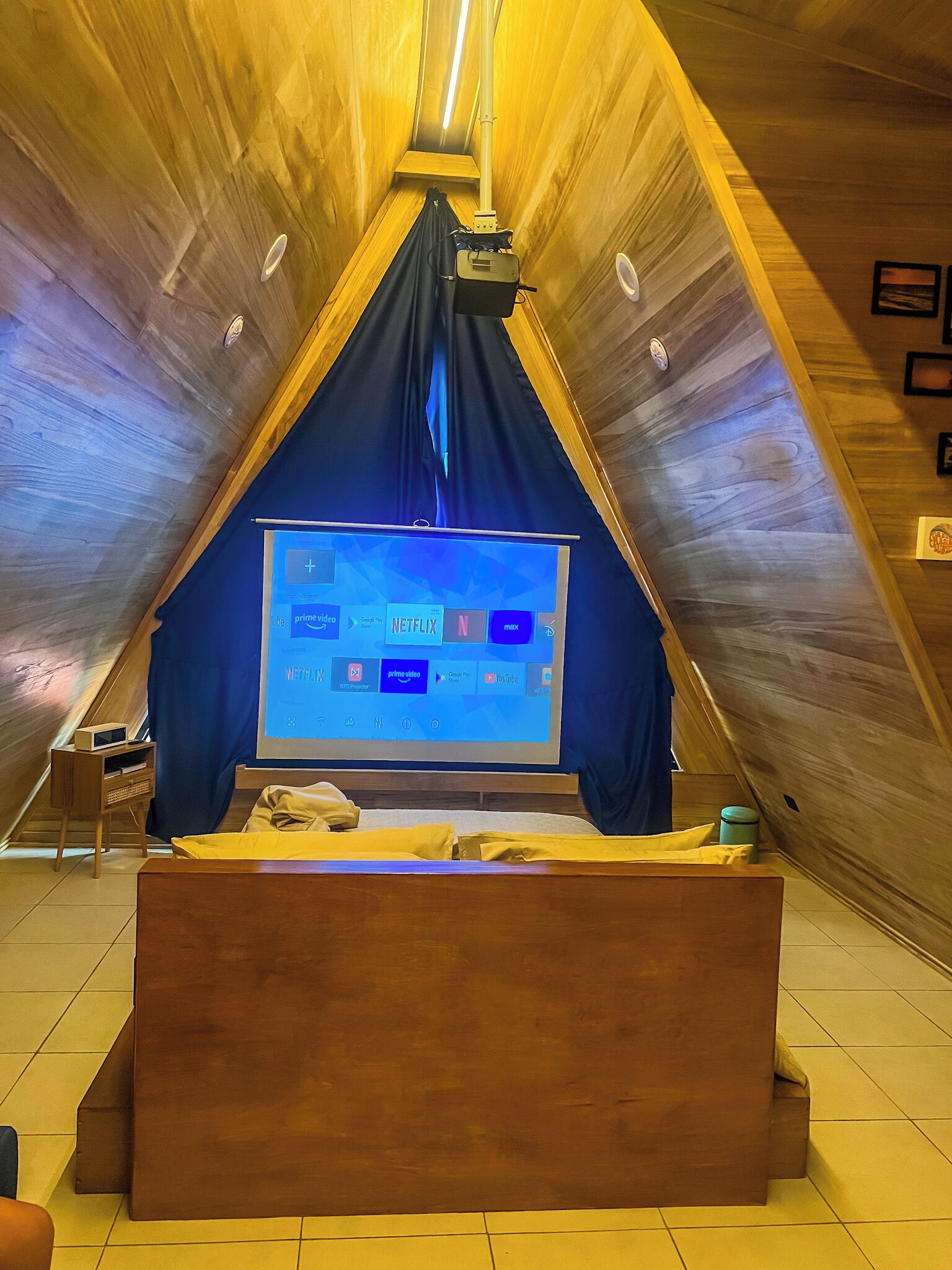A cozy attic room with a large TV screen displaying streaming services, a wooden bed, and a small side table.