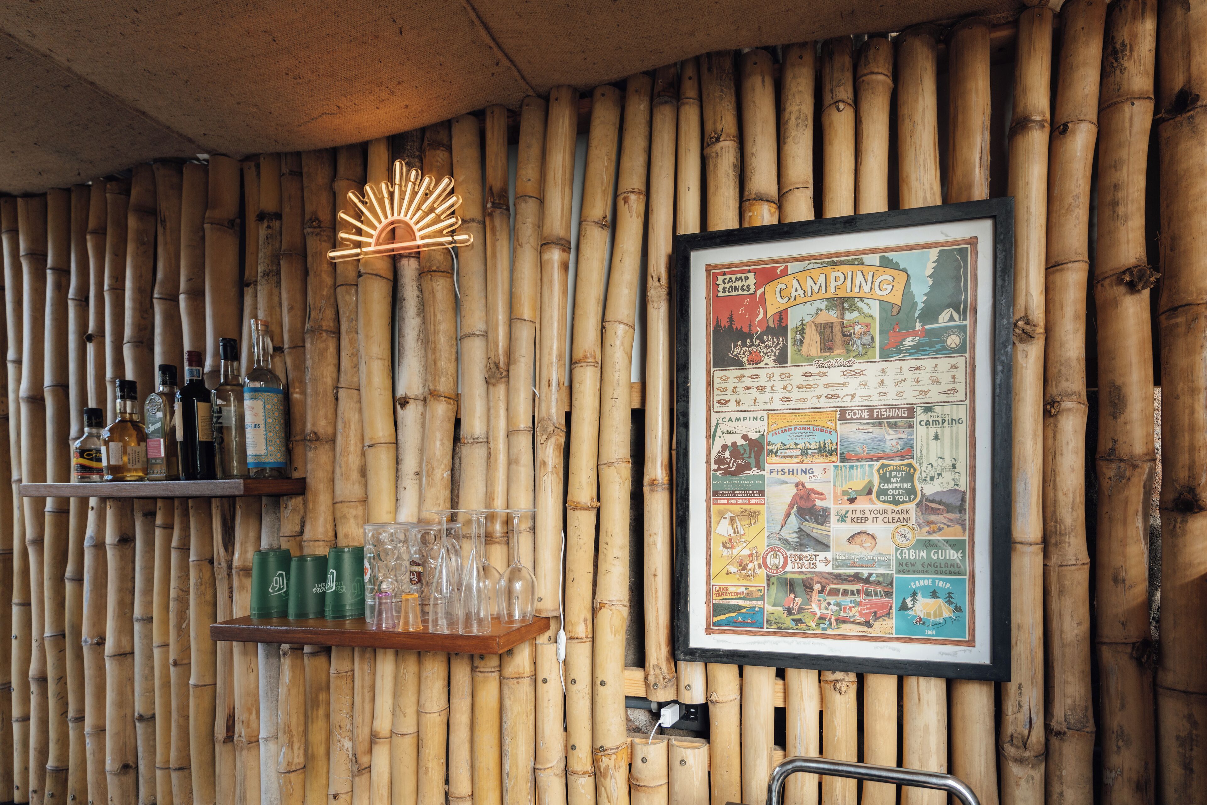 A bamboo wall with a framed camping poster, shelves with bottles and glasses, and a neon sign.