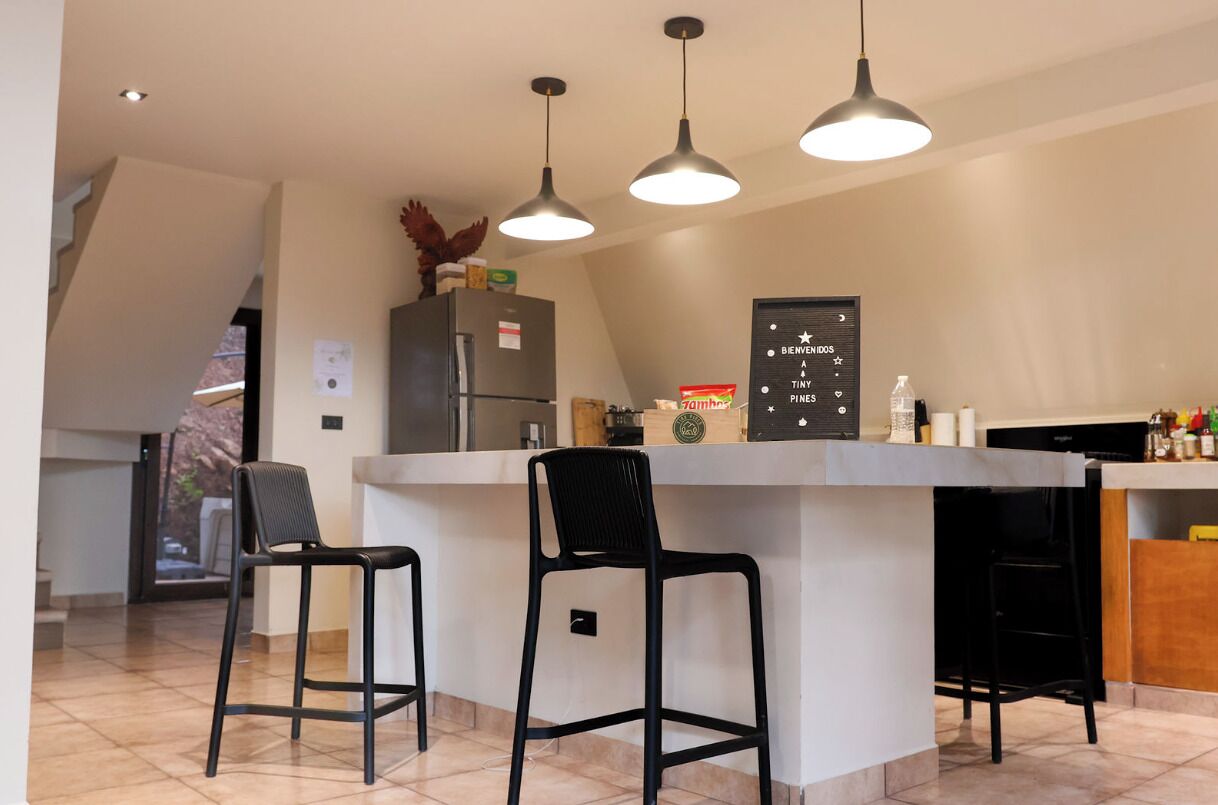 Una cocina con isla central, taburetes de bar, iluminación colgante y un refrigerador.