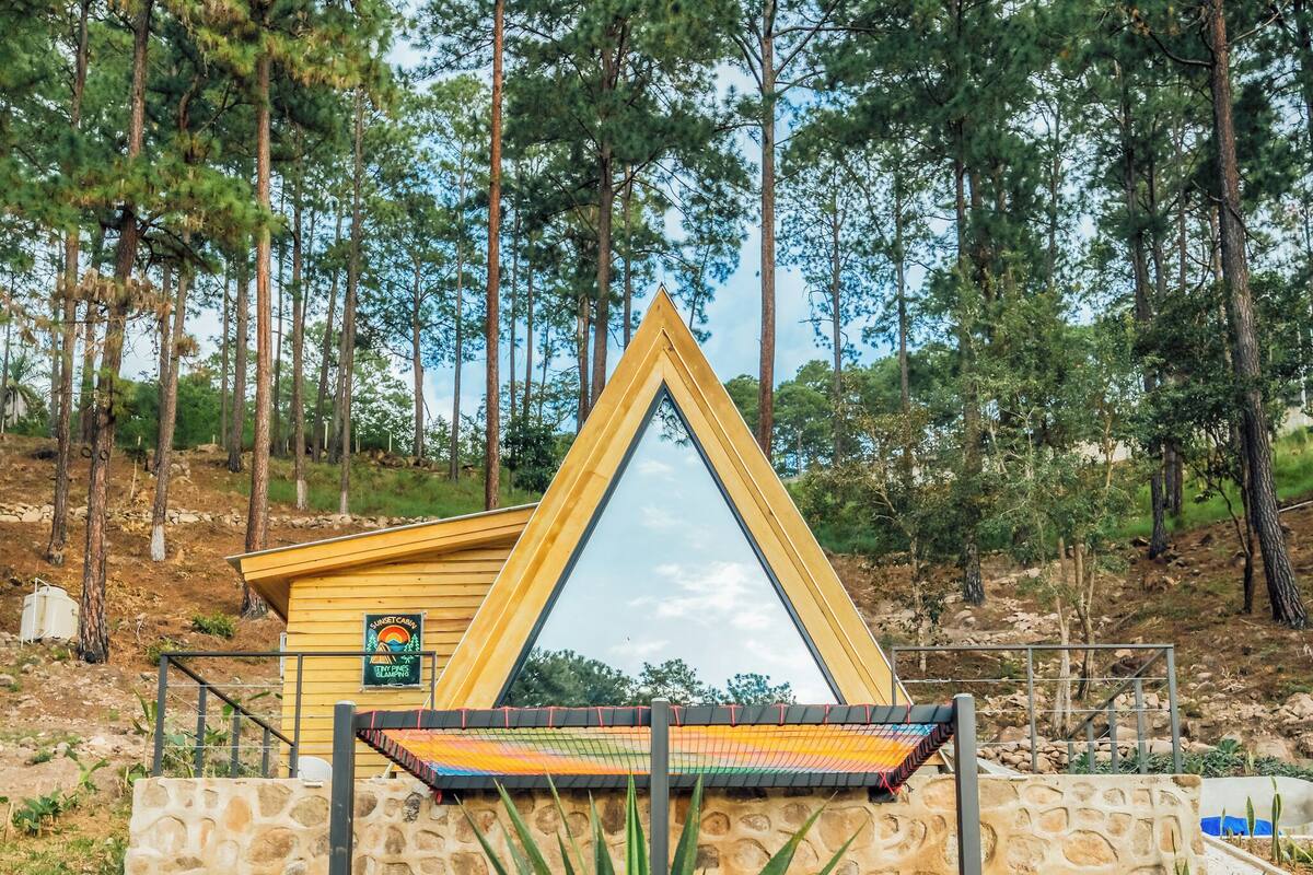 A triangular wooden structure with a glass roof, surrounded by a stone wall and a hammock.
