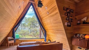 A cozy attic bedroom with a sloped ceiling, a large bed, a small desk, and a window overlooking a forest.