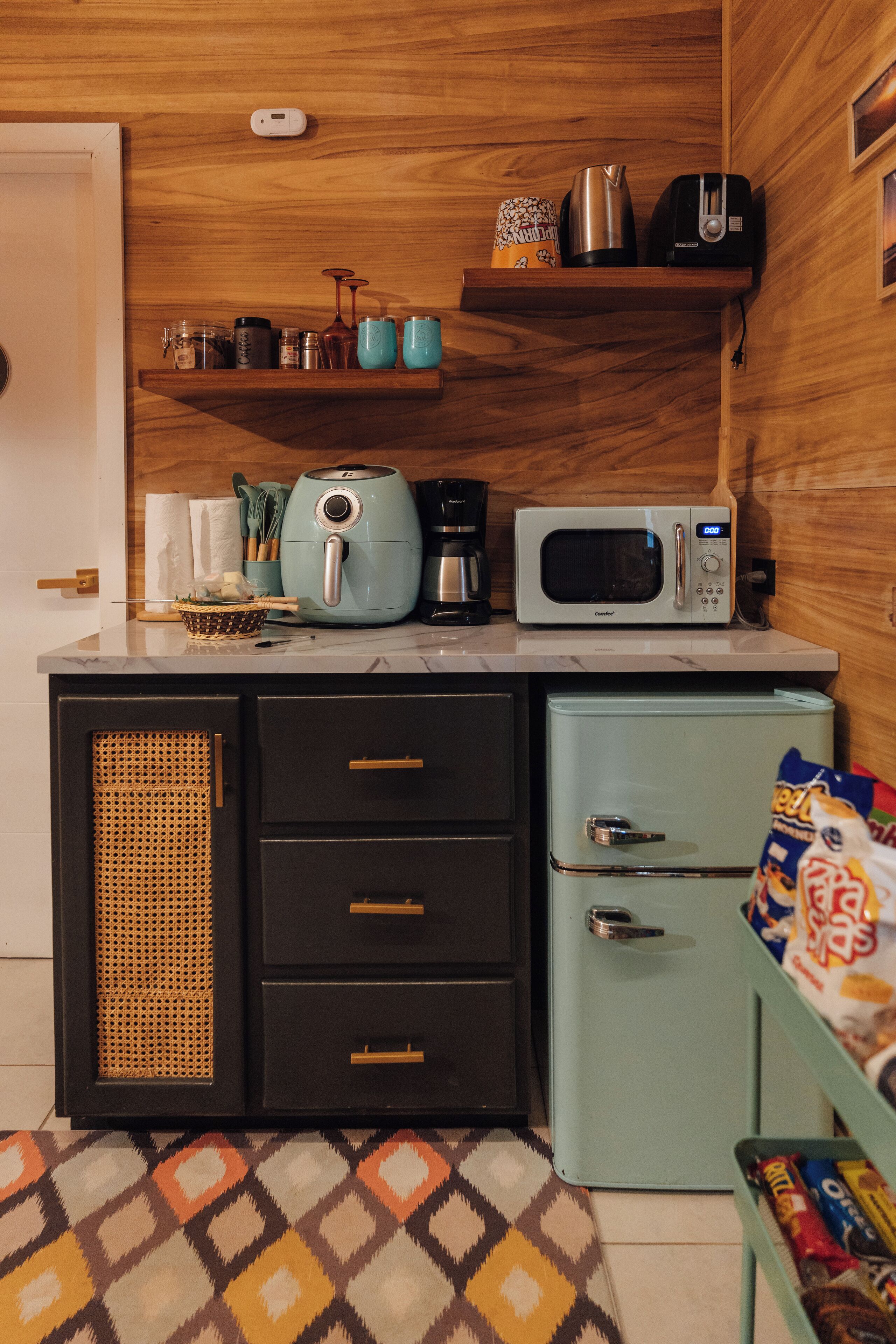 A compact kitchen with a microwave, toaster, and coffee maker on the countertop, a small refrigerator, and a shelf with kitchen items.