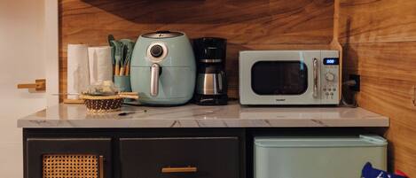 A compact kitchen with a microwave, toaster, and coffee maker on the countertop, a small refrigerator, and a shelf with kitchen items.