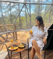Uma pessoa sentada à mesa com uma refeição e bebida, dentro de uma estrutura tipo tenda com vista para árvores.