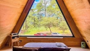 A triangular wooden room with a bed, a small table, and a window offering a view of trees and a car.