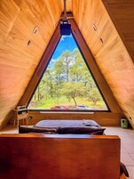 Quarto de madeira em formato triangular com uma cama, uma mesinha e uma janela com vista para árvores e um carro.