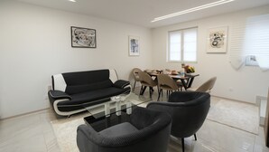A modern living room with a black sofa, glass coffee table, and black chairs. There is a dining area with a table and chairs, and artwork on the walls. - Le Marbre Blanc 4* - Les Suites du Lion (Rosheim)
