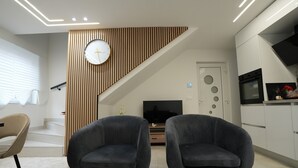 A modern living room with two dark armchairs, a glass coffee table, a wooden sloped ceiling, a TV, and a kitchen area with white cabinets and a black oven. - Le Marbre Blanc 4* - Les Suites du Lion (Rosheim)