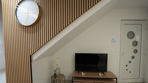 A modern living room with a wooden sloped ceiling, a flat-screen TV on a wooden TV stand, a small side table with a vase of flowers, and a door with circular cutouts. - Le Marbre Blanc 4* - Les Suites du Lion (Rosheim)