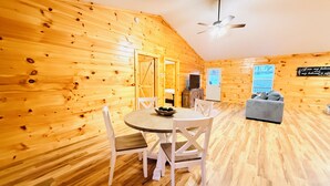 Interior acogedor de una cabaña con paredes de madera, un comedor con mesa y sillas, un sofá gris y un ventilador de techo.
