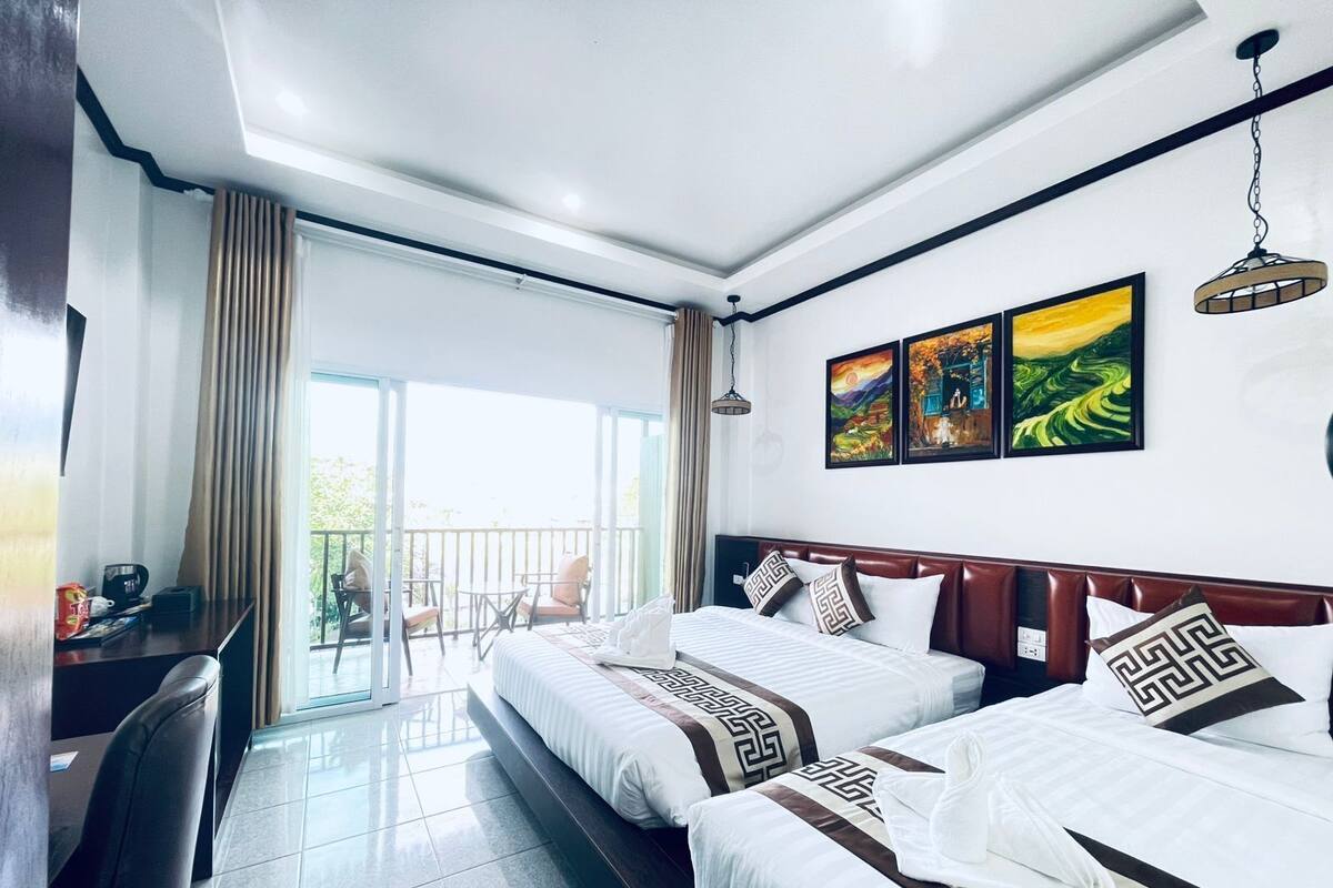 A hotel room with two beds, a desk, and a balcony with chairs.