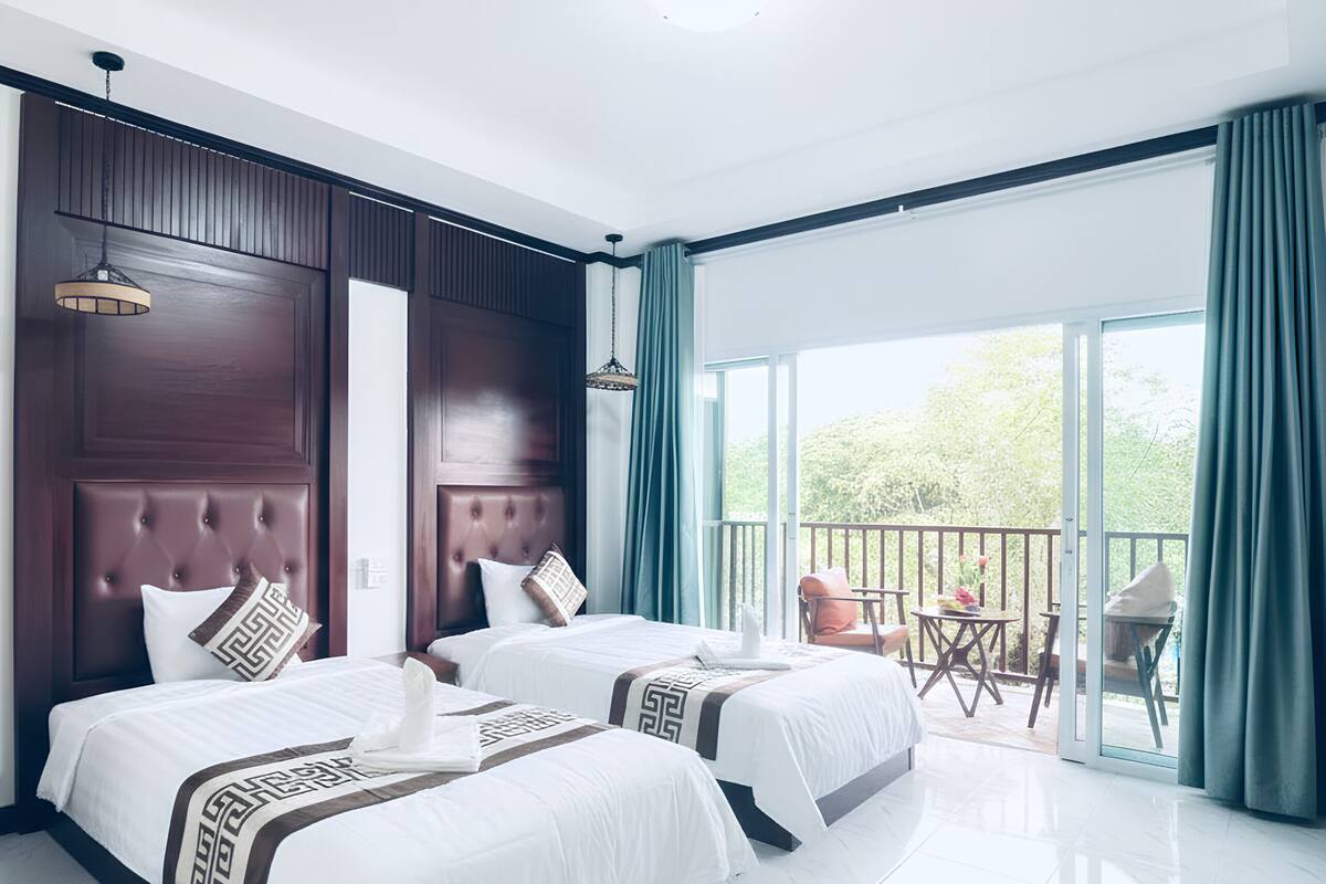 A hotel room with two beds, a balcony, and a view of greenery.