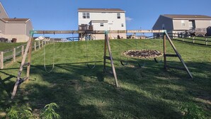 A grassy area with a swing set and a wooden play structure. In the background, there are two houses with a clear blue sky above.