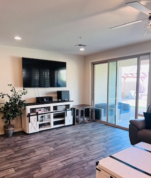 A living room with a flat-screen TV, a white entertainment unit, a potted plant, and a sliding glass door leading to an outdoor area. - Cozy Desert Winter Haven (Peoria)